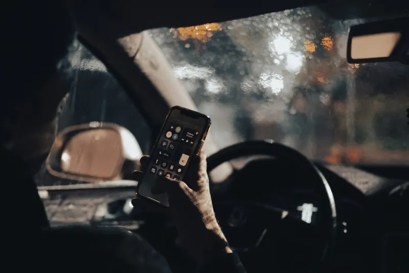 Symbolbild: Hand mit Smartphone am Lenkrad eines Autos bei Nacht und Regen
